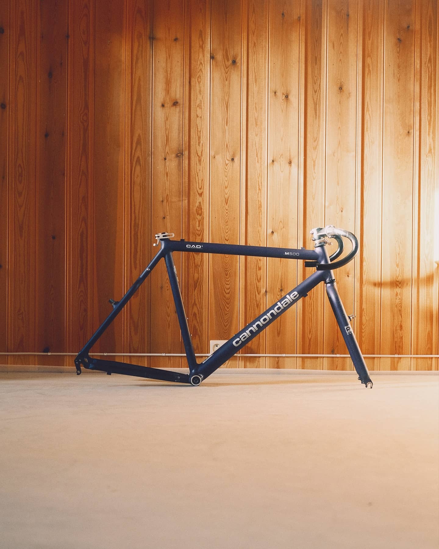 A bare navy Cannondale CAD3 M500 frame and fork standing on a concrete floor against a pine-panelled wall, the Cannondale name in white down the down tube.
