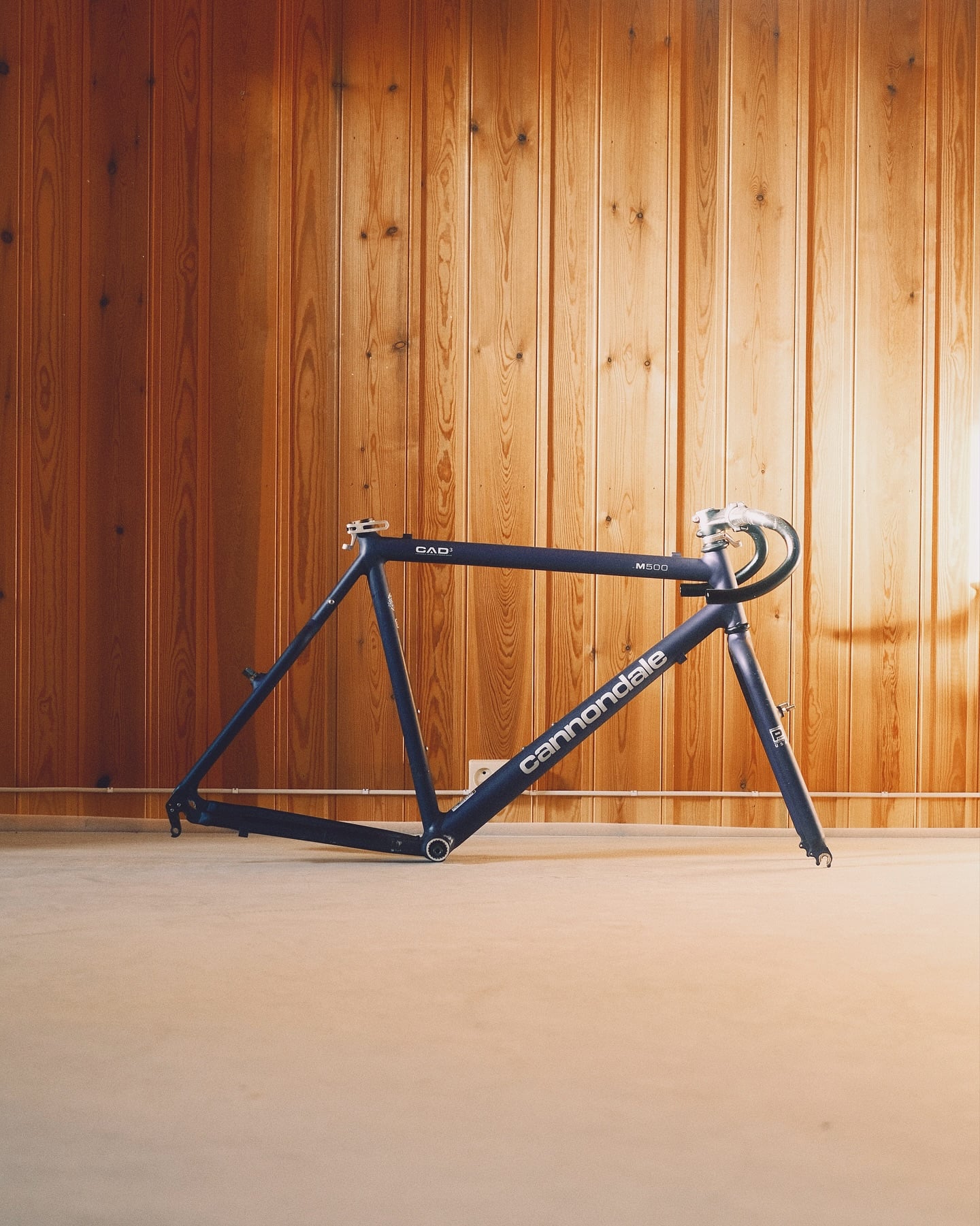 A bare navy Cannondale CAD3 M500 frame and fork standing on a concrete floor against a pine-panelled wall, the Cannondale name in white down the down tube.