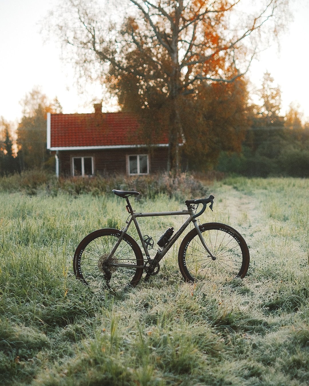 A dark Cannondale cyclocross bike with a frame bag, standing on a frosted meadow track with a red-and-white cottage and autumn trees behind.
