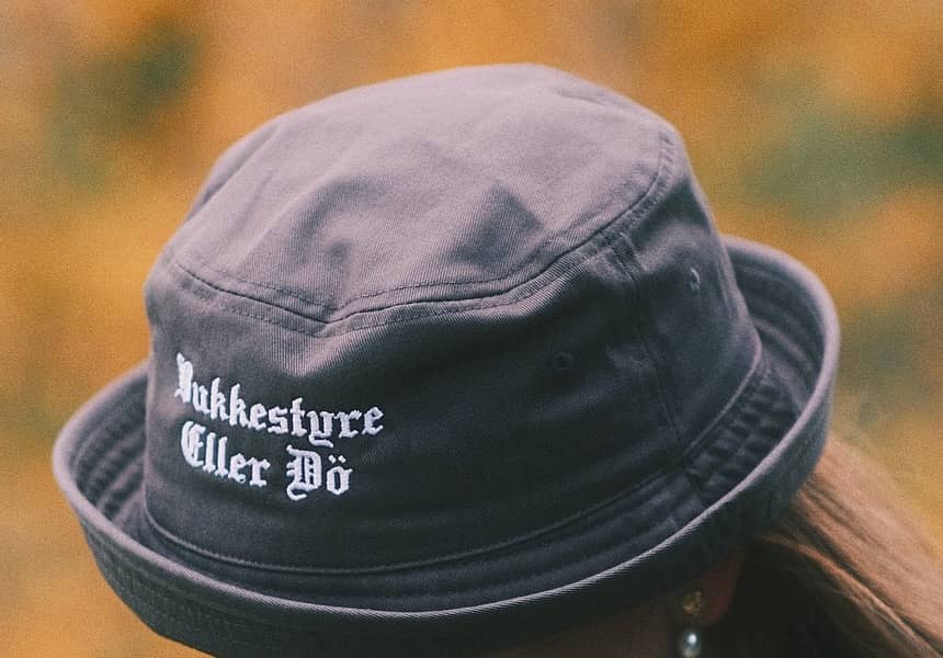 A dark bucket hat embroidered in white blackletter with the words Jukkestyre Eller Dö, worn against blurred autumn foliage.