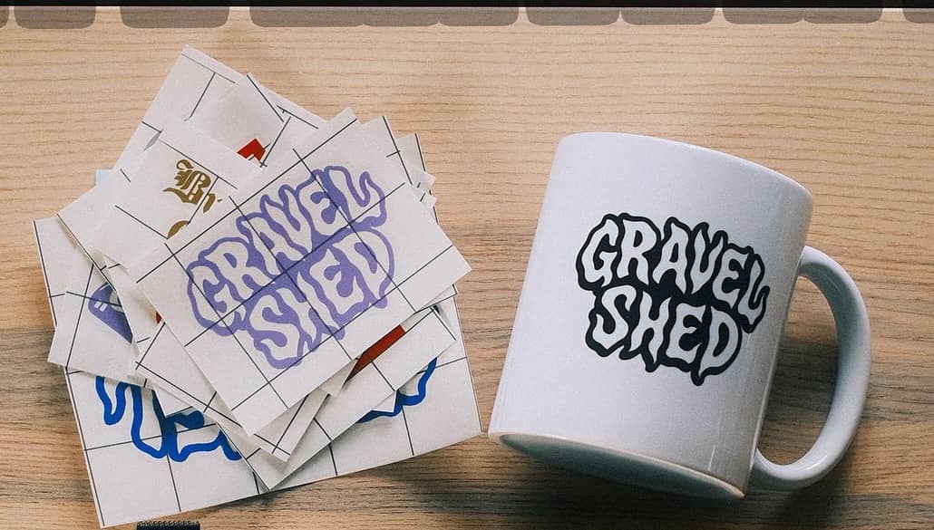 A stack of Gravel Shed wordmark stickers in purple, blue and gold beside a white enamel mug with the same wordmark, on a wooden table.