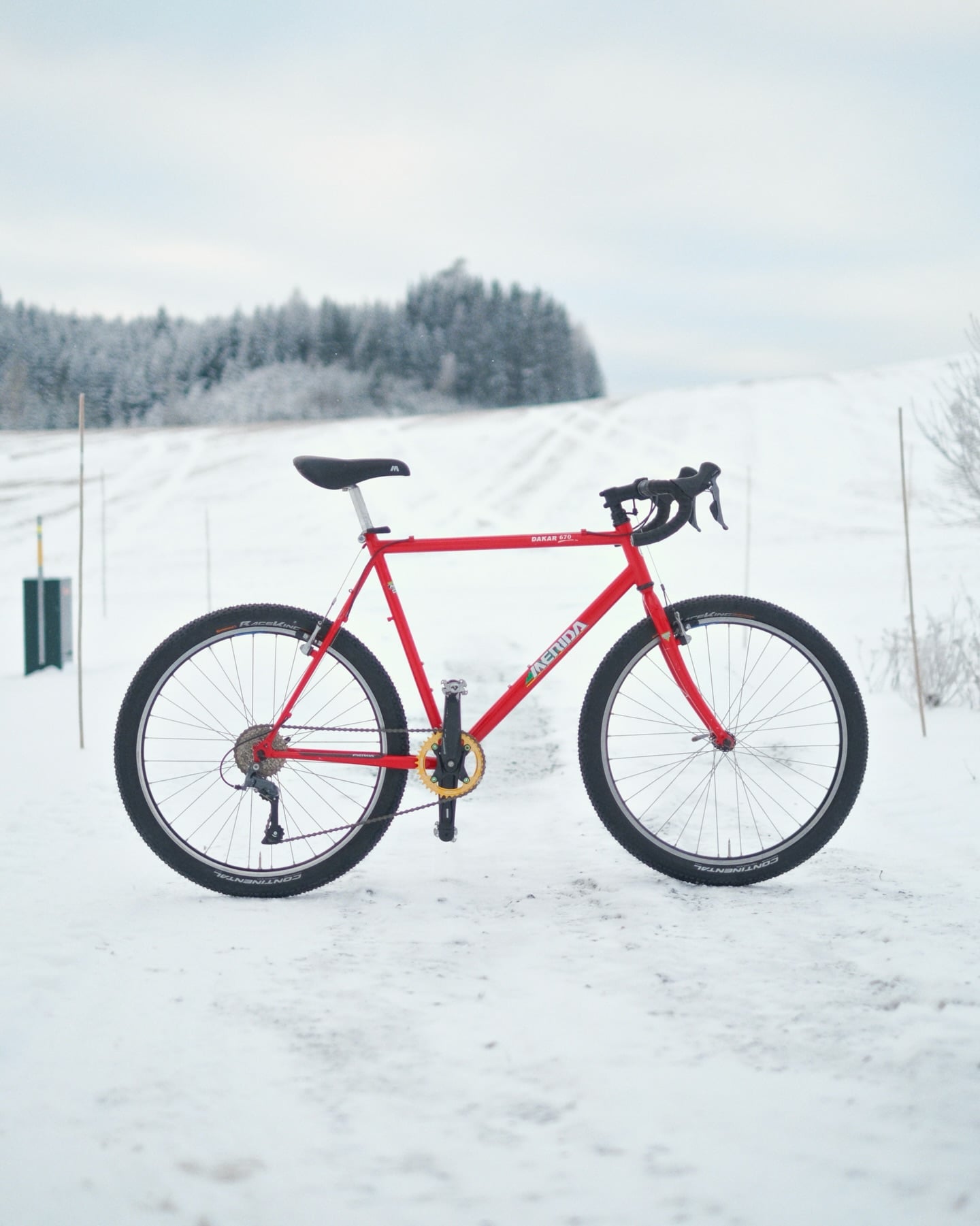 A bright red Merida Dakar 670 with drop bars and a gold chainring, standing on a snow-covered track with frosted forest behind.