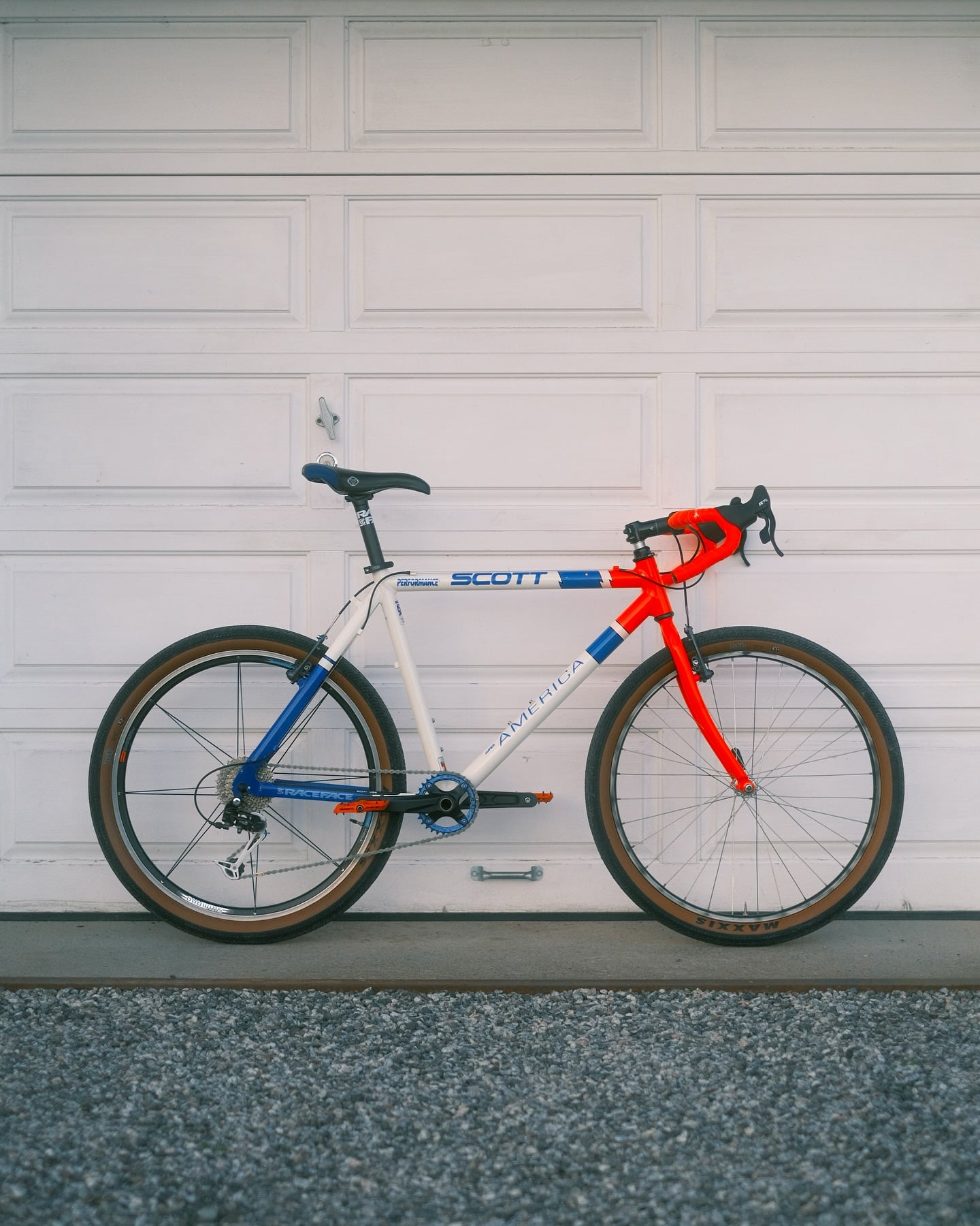A white Scott with blue and red panels, red drop-bar tape and tan-wall knobby tyres, standing on gravel against a white garage door.