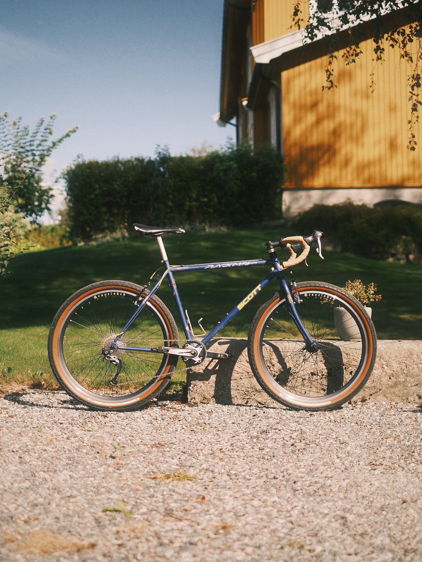 A navy Scott Racing with pale bar tape and tan-wall tyres, standing on gravel in front of a yellow timber barn.