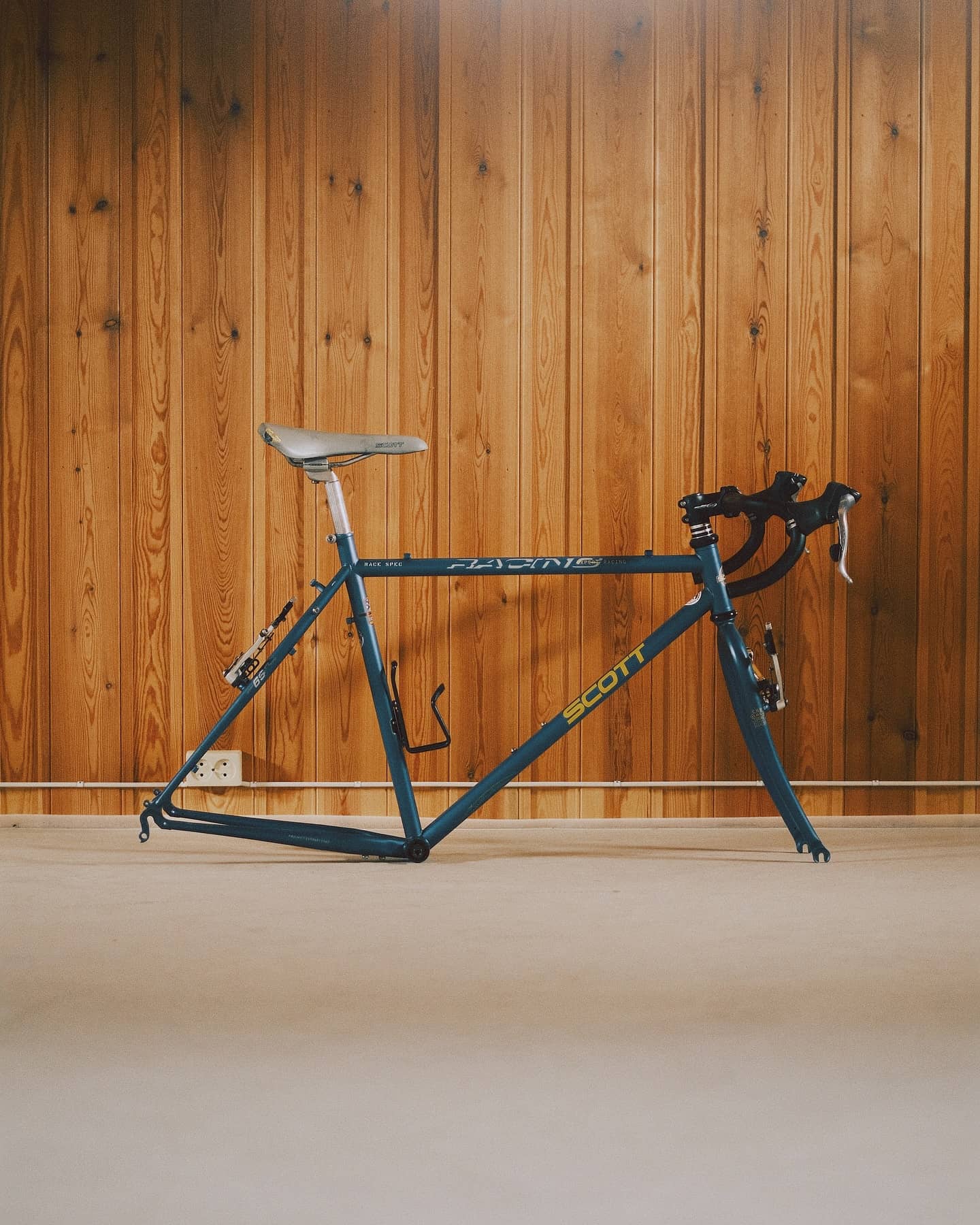 A bare teal Scott Racing frame and fork with saddle and stem fitted but no wheels, leaning against a pine-panelled workshop wall.